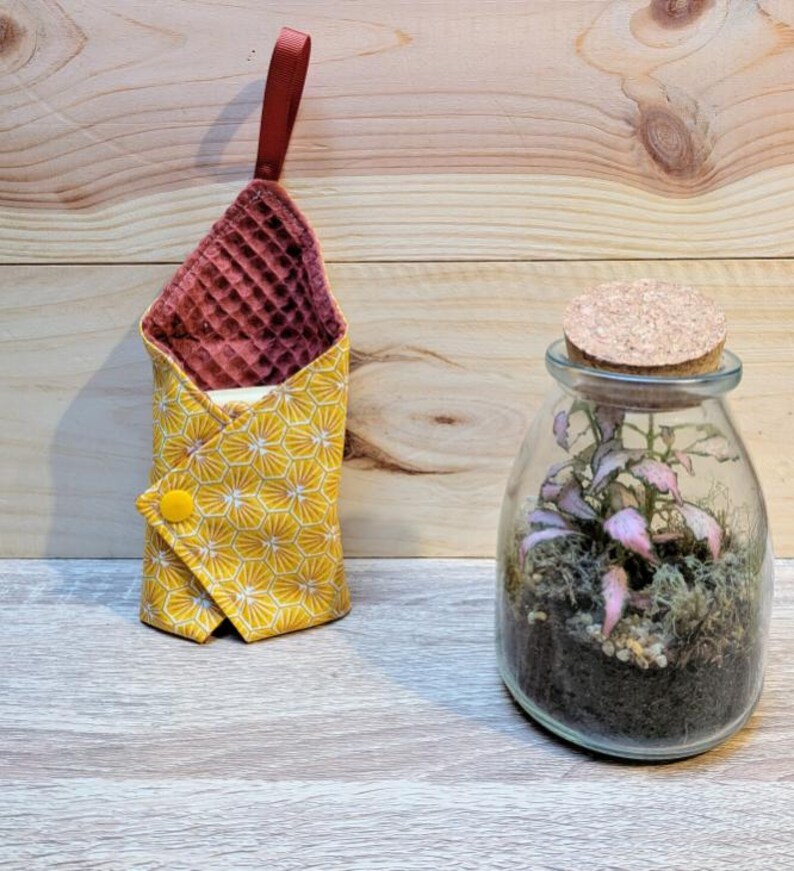 Puede incluir: Una bolsa de tela amarilla con un patr&oacute;n de panal y un cierre de bot&oacute;n amarillo, junto a un terrario de vidrio con tapa de corcho. La bolsa tiene un interior burdeos y un lazo burdeos.