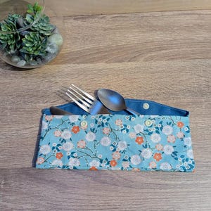 Può includere: Un portaposate con stampa floreale blu contenente una forchetta, un cucchiaio e un coltello. Il portaposate in tessuto presenta uno sfondo azzurro con un motivo di fiori bianchi e arancioni. È fissato con due piccoli bottoni.