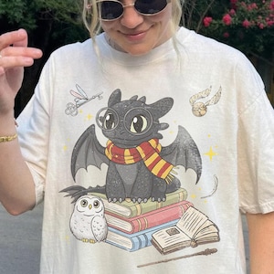 Op de afbeelding: Een crèmekleurig T-shirt met korte mouwen en een afbeelding van een zwarte draak met een ronde bril en een rood-geel gestreepte sjaal. De draak zit op een stapel boeken naast een witte uil, een vliegende sleutel, een gouden snaai en een toverstaf.