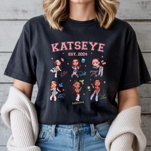 Peut inclure: T-shirt noir avec l'inscription rose "KATSEYE EST. 2024" et des illustrations de filles de dessins animés. Chaque fille a une coiffure et une tenue différentes. Le t-shirt est porté par une personne.