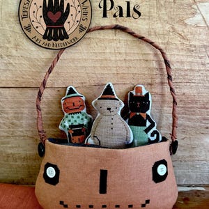Può includere: Un cestino in tessuto color pesca con un volto di zucca ricamato e due occhi a bottone bianchi. All'interno ci sono tre figure a punto croce: una zucca, un orso e un gatto nero. Il testo riporta Pumpkin Pals, XS371 e Teresa Kogut.