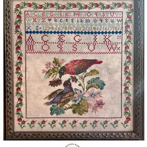 Op de afbeelding: Een ingelijste kruissteek sampler met het woord Beloved bovenaan, een alfabet, cijfers en een centraal ontwerp van twee vogels op bloeiende takken. Het werk is omrand door een herhalend patroon van kleine rode bessen en groene bladeren.