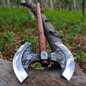 May include: A double-bladed axe with a polished steel head and a wooden handle. The axe head features decorative etched designs. The handle is a dark brown color. The axe is leaning against a tree branch.