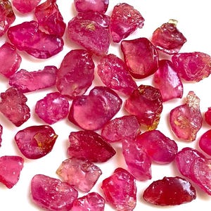 Może przedstawiać: Kolekcja surowych, nieoszlifowanych kamieni szlachetnych rubinu. Kamienie różnią się rozmiarem i kształtem, prezentując gamę odcieni różu i czerwieni. Kamienie są rozrzucone na białej powierzchni, ukazując ich naturalne, nieregularne formy.