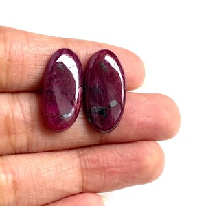 May include: Two oval-shaped, polished ruby gemstones. The rubies are a deep, rich red colour with dark inclusions. The gemstones are held in a hand against a white background. Suitable for jewellery making.