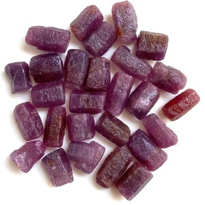 Puede incluir: Una colección de piedras preciosas de rubí en bruto, sin cortar. Las piedras varían en tonos de púrpura intenso y marrón rojizo, con una superficie rugosa y texturizada. Las piedras están sobre un fondo blanco.