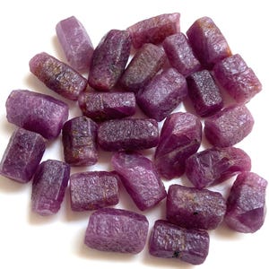 Könnte beinhalten: Eine Ansammlung roher, ungeschliffener Rubin-Edelsteine. Die Steine variieren in Größe und Form und weisen einen tiefen rot-violetten Farbton auf. Die raue Textur und die natürlichen Formen der Rubine sind vor weißem Hintergrund sichtbar.