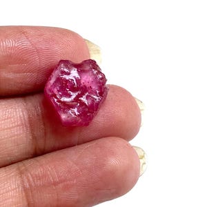 May include: A rough-cut, natural ruby gemstone. The ruby is a deep, vibrant pinkish-red color with a slightly translucent appearance. The gemstone has an irregular, textured surface and is held in a hand against a white background.