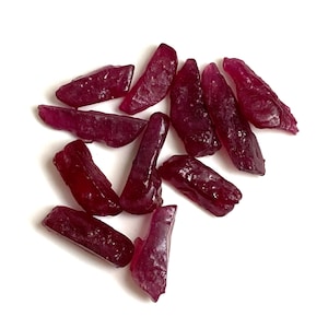 Può includere: Una collezione di gemme di rubino grezze, non tagliate. Le gemme sono di un colore rosso intenso e ricco, con un aspetto leggermente traslucido. Le forme sono irregolari, con bordi frastagliati e dimensioni variabili. I rubini sono sparsi su uno sfondo bianco.