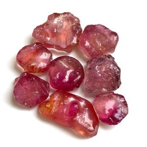 Könnte beinhalten: Eine Sammlung roher, ungeschliffener Edelsteine in Rosa- und Rottönen. Die Steine variieren in Größe und Form und weisen eine durchscheinende Qualität auf. Die Edelsteine sind auf weißem Hintergrund verstreut, was ihre natürlichen, organischen Formen hervorhebt.