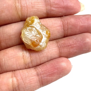 Puede incluir: Una gema en bruto, sin cortar, de color amarillo dorado con apariencia translúcida. La piedra tiene una forma irregular y una textura ligeramente rugosa. La gema se sostiene en una mano, con un fondo blanco.