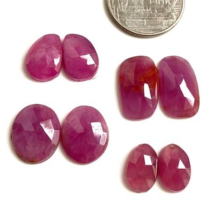 Pode incluir: Várias pedras preciosas de rubi facetadas, em formas ovais e de gota, em tamanhos variados, são exibidas sobre um fundo branco. Os rubis exibem um tom vermelho profundo e são colocados perto de uma moeda de prata.