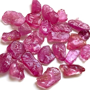 Könnte beinhalten: Eine Sammlung roher, ungeschliffener Rubin-Edelsteine. Die Steine variieren in Größe und Form und weisen eine Reihe von rosa und roten Farbtönen auf. Die Edelsteine haben eine raue, natürliche Textur und eine durchscheinende Qualität.