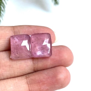 May include: Two square, pink gemstones with white lightning bolt-shaped inclusions are held in a hand. The gemstones have a slightly translucent appearance and a polished surface. The background is white.