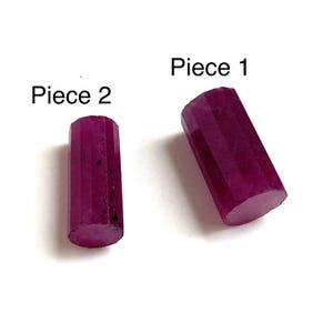 May include: Two faceted, cylindrical ruby gemstones are displayed against a white background. The gemstones are a deep, rich magenta color. The text "Piece 1" and "Piece 2" are above each gemstone.
