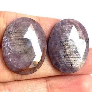 Peut inclure: Deux pierres précieuses ovales avec une taille à facettes. Les gemmes sont d'une couleur violette sourde avec des stries et des inclusions internes. Les pierres sont tenues dans une main, sur fond blanc.
