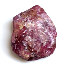 May include: A raw, natural gemstone in a deep magenta colour, with lighter pink and beige areas. The stone has a textured surface and an irregular shape, set against a clean white background. Suitable for jewellery making or collecting.