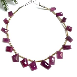Peut inclure: Un collier composé de pierres précieuses rubis rectangulaires et facettées, enfilées sur un fil doré. Les rubis sont d'un rose profond et riche. Le collier est présenté sur un fond blanc.