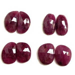 May include: Four pairs of faceted, oval-shaped ruby gemstones. The rubies are a deep, rich red color with visible facets that catch the light. Each pair is displayed against a plain white background, showcasing the gemstones' natural beauty.