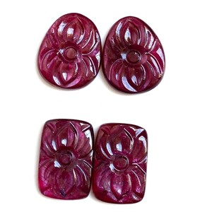 Könnte beinhalten: Vier geschnitzte Rubin-Edelsteine. Zwei sind oval und zwei rechteckig. Jeder Edelstein weist ein detailliertes Blumendesign auf, das in die Oberfläche geschnitzt ist. Die Rubine sind tiefrot, und der Hintergrund ist weiß.