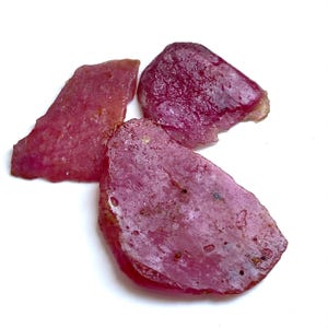 Puede incluir: Tres piedras preciosas de rubí en bruto, de un tono rojo-rosado intenso, se muestran sobre un fondo blanco. Las piedras tienen formas irregulares y una superficie texturizada, mostrando variaciones naturales.