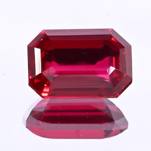 May include: A faceted, rectangular-cut ruby gemstone. The gem is a deep, saturated red color with a high degree of clarity. The stone is displayed on a reflective surface, showing its mirrored image.