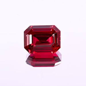 May include: A faceted, rectangular-cut, deep red gemstone. The gem has a reflective surface and is set against a white background. The cut of the gem creates multiple facets, enhancing its brilliance and color.