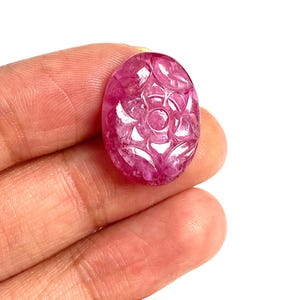 May include: A carved, oval-shaped gemstone in shades of pink and red. The gemstone features a floral design carved into its surface. The stone is held in a hand against a white background. The gemstone is approximately 2.5 cm in length.