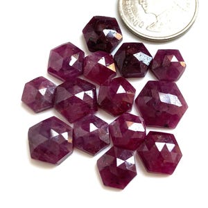 Puede incluir: Un conjunto de gemas facetadas, de forma hexagonal y de color rojo intenso. Las gemas están dispersas sobre una superficie blanca, con una moneda de plata en el fondo. Las gemas tienen múltiples facetas que reflejan la luz. Adecuado para la creación de joyas.