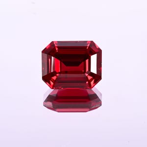 May include: A faceted, rectangular-cut, deep red gemstone. The gem is highly reflective, with a polished surface and sharp edges. The gem is set against a white background, with its reflection visible below.