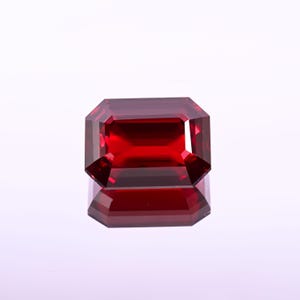 May include: A faceted, rectangular-cut, deep red gemstone. The gem is highly reflective, with sharp edges and a polished surface. The stone is set against a white background, with its reflection visible below.