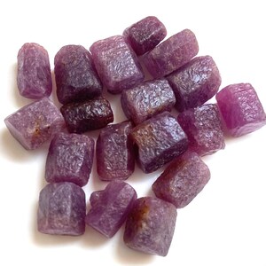 May include: A collection of raw, uncut ruby gemstones. The stones vary in size and shape, displaying a deep reddish-purple hue. The rough texture and natural form of the rubies are visible against a white background, highlighting their organic beauty.