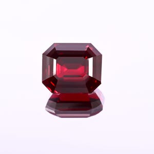 May include: A faceted, rectangular-cut, deep red gemstone. The gem has a reflective surface, and its facets catch the light, creating a brilliant display. The gem is set against a white background, with its reflection visible below.