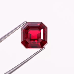 May include: A faceted, rectangular-cut, deep red gemstone is held by a pair of silver tweezers against a white background. The gemstone has a square shape with beveled edges and a brilliant, reflective surface.