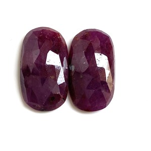 Peut inclure: Deux pierres précieuses ovales et facettées d'un rouge-violet profond. Les gemmes ont une surface polie et réfléchissante, présentant un motif complexe de facettes internes. Les pierres sont posées sur un fond blanc.