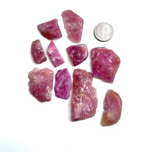 Puede incluir: Una colección de piedras de turmalina rosa en bruto, de varios tamaños y formas, exhibidas sobre una superficie blanca. Las piedras muestran una gama de tonos rosados, desde claros hasta magenta intenso, con texturas visibles y formaciones naturales. Se incluye una moneda como referencia de tamaño.