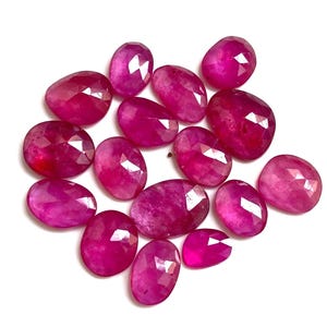 Puede incluir: Una colección de gemas de rubí facetadas, de forma ovalada y de varios tamaños. Las gemas son de un color rojo-rosado intenso y vibrante, dispuestas sobre un fondo blanco. Las gemas tienen una superficie pulida y reflectante.