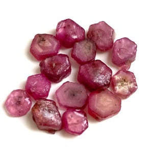 Könnte beinhalten: Eine Ansammlung roher, ungeschliffener Rubin-Edelsteine. Die Steine variieren in Form und Größe und weisen eine tiefrosa bis rote Farbe auf. Einige Steine haben eine durchscheinende Qualität, die innere Strukturen offenbart. Die Edelsteine liegen auf weißem Grund.