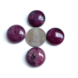 Op de afbeelding: Vier gepolijste, ronde, diep magenta edelstenen met donkere insluitsels zijn gerangschikt rond een zilveren munt. De munt heeft tekst en een ontwerp in het midden. De edelstenen hebben een glad, reflecterend oppervlak en staan tegen een witte achtergrond.
