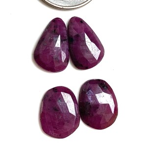 Peut inclure: Quatre pierres précieuses rouge foncé, taillées en forme ovale, avec une pièce de monnaie en arrière-plan. Les pierres ont une surface polie et réfléchissante, sur fond blanc. Il s'agit probablement de rubis.