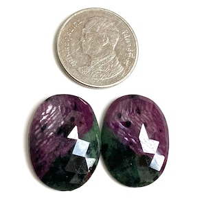 May include: Two oval-shaped gemstones with a faceted cut, displaying a mix of deep red and green hues. A silver coin is positioned above the gemstones. The gemstones are set against a white background.