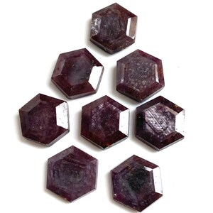 May include: Seven faceted, hexagonal gemstones in a deep burgundy color are arranged on a white background. The gems have a polished appearance, with visible internal patterns and reflective surfaces. The arrangement suggests a collection of precious stones.
