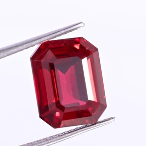 Peut inclure: Une pierre précieuse rouge foncé, taillée en rectangle, tenue par une pince argentée. La gemme a une surface polie et vibrante et une forme symétrique, reflétant la lumière. Il s'agit d'un rubis.