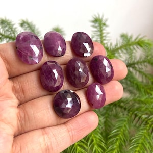 Könnte beinhalten: Acht facettierte, ovale Rubine in verschiedenen Größen. Die tiefroten bis violetten Edelsteine werden in einer Hand gehalten, vor einem Hintergrund aus grünem Blattwerk.