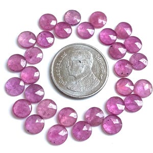 Può includere: Una collezione di gemme rosa rotonde e sfaccettate disposte attorno a una moneta d'argento. Le gemme hanno un aspetto lucido e sono di una vibrante tonalità rosa. La moneta presenta un ritratto e del testo.