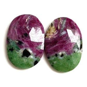 Może przedstawiać: Dwa owalne kamienie szlachetne o fasetowanej powierzchni. Kamienie charakteryzują się żywą paletą barw, z mieszanką głębokiej magenty, zieleni i czerni. Kamienie szlachetne są prawdopodobnie używane do wyrobu biżuterii.