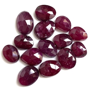 May include: A collection of faceted, oval-shaped ruby gemstones. The rubies are a deep, rich red color with multiple facets that catch the light. The gemstones are scattered across a white surface.