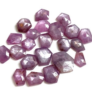 Può includere: Una collezione di pietre preziose sfaccettate, di forma irregolare, in varie tonalità di viola. Le pietre preziose hanno una superficie lucida e riflettente, sparse su uno sfondo bianco. Le gemme sono probabilmente utilizzate per la creazione di gioielli.
