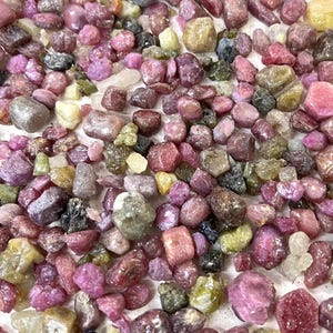 Puede incluir: Primer plano de una colección de pequeñas piedras preciosas pulidas en tonos rosa, morado, verde y amarillo. Las piedras, de diferentes tamaños y formas, crean una exhibición colorida y texturizada. Ideal para la creación de joyas o coleccionar.