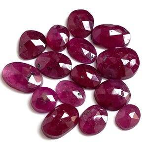 Könnte beinhalten: Eine Ansammlung facettierter Rubine in ovalen und runden Formen. Die Rubine weisen eine tiefe, satte rote Farbe mit reflektierenden Facetten auf. Die Edelsteine liegen auf einem weißen Hintergrund, was ihre Eignung für Schmuckherstellung unterstreicht.